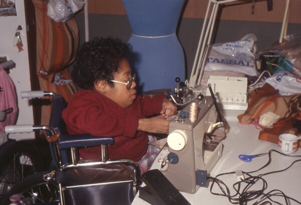 Image 1: Digitised slide of Het Dorp resident Josje Matser in a wheelchair sitting at a table at the Sewing Ring, working on a sewing machine. Date unknown [1977-1985]. Property of Siza.