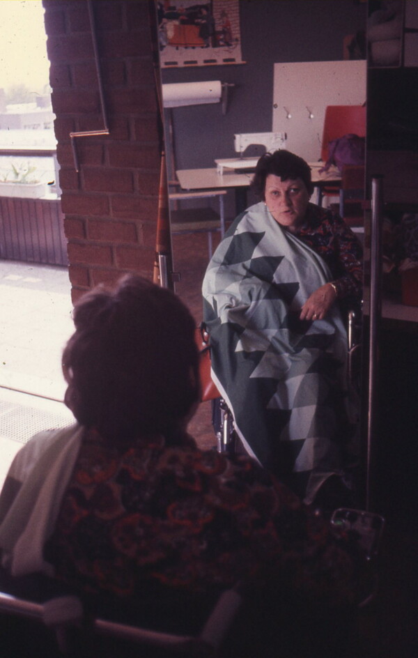 Image 3: Digitised slide of Het Dorp resident Wil Goos in a wheelchair sitting in front of a full length mirror at the Sewing Ring, a piece of fabric draped across her body. Date unknown [1977-1985]. Property of Siza.